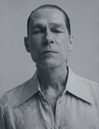Portrait of an adult man with slicked back hair wearing a patterned button-down shirt against a neutral backdrop.