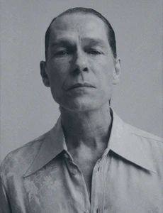Portrait of an adult man with slicked back hair wearing a patterned button-down shirt against a neutral backdrop.