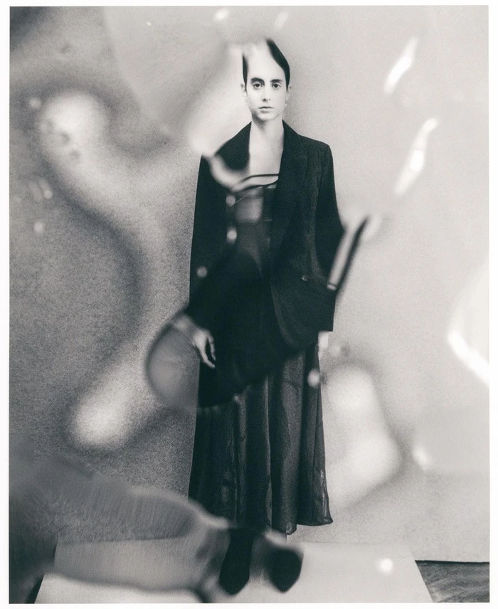 Monochrome portrait of a person in a dark blazer and skirt viewed through a distorted glass surface