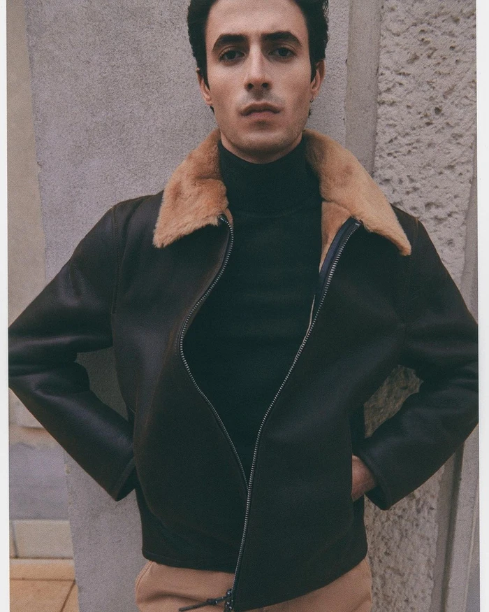 Portrait of a man in a dark brown shearling collar jacket and black turtleneck standing against a textured stone wall.