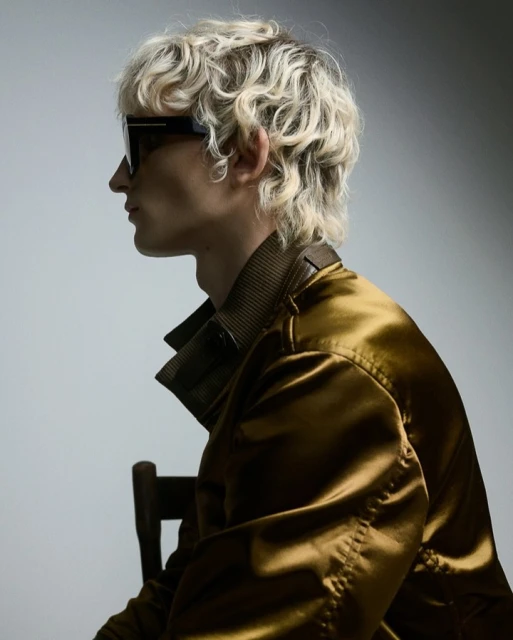 Profile shot of a young adult male with blonde curly hair wearing sunglasses and a golden jacket sitting against a light backdrop.