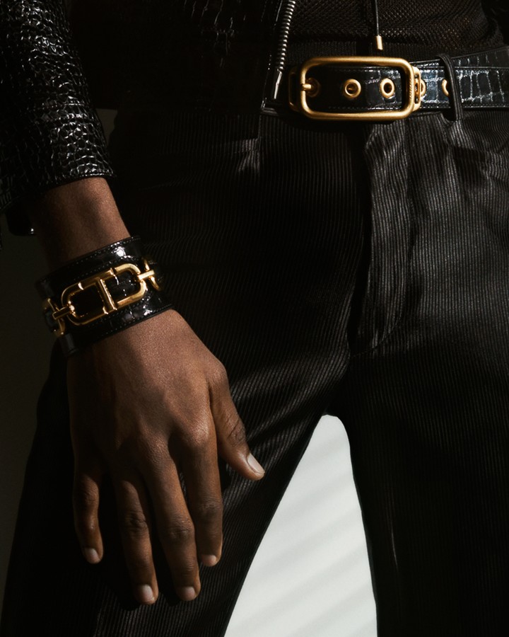 Cropped shot of a black model's hand wearing a black and gold bracelet, black belt, and black pants in a studio setting.