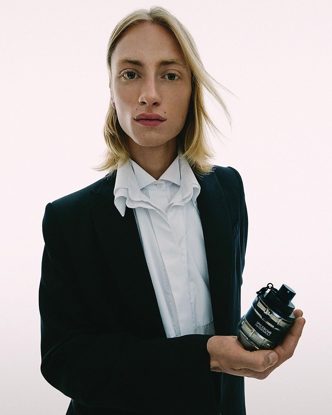 Young adult man in a black suit holding a silver cologne bottle. Shot in a bright photo studio with simple and clean aesthetic.