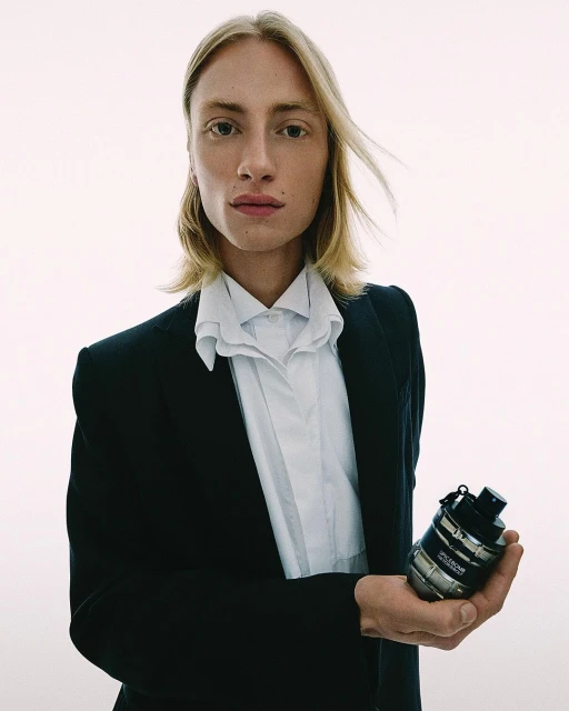 Young adult man in a black suit holding a silver cologne bottle. Shot in a bright photo studio with simple and clean aesthetic.
