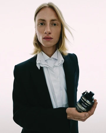Young adult man in a black suit holding a silver cologne bottle. Shot in a bright photo studio with simple and clean aesthetic.