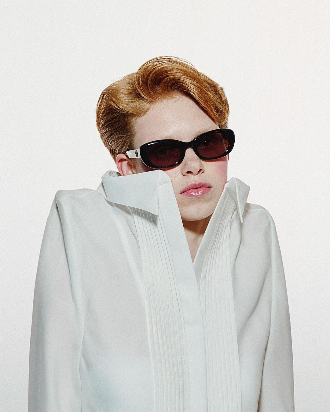 Model with red hair wearing dark sunglasses and a structured white shirt against a plain background
