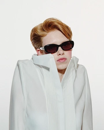Model with red hair wearing dark sunglasses and a structured white shirt against a plain background