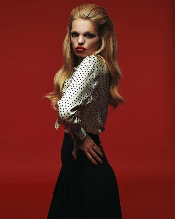 Woman in polka dot blouse and dark skirt standing against a solid red backdrop by Ferry Van Der Nat