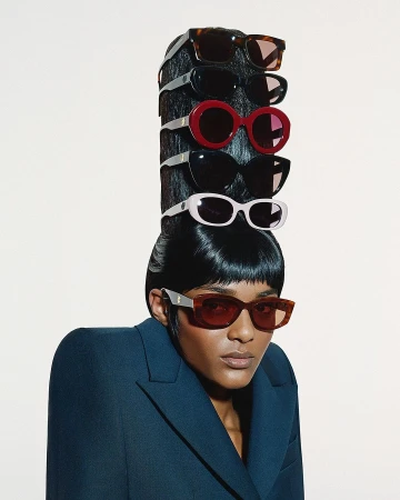 Model wearing dark sunglasses with multiple stacked pairs mounted vertically in an elaborate tall black hair structure