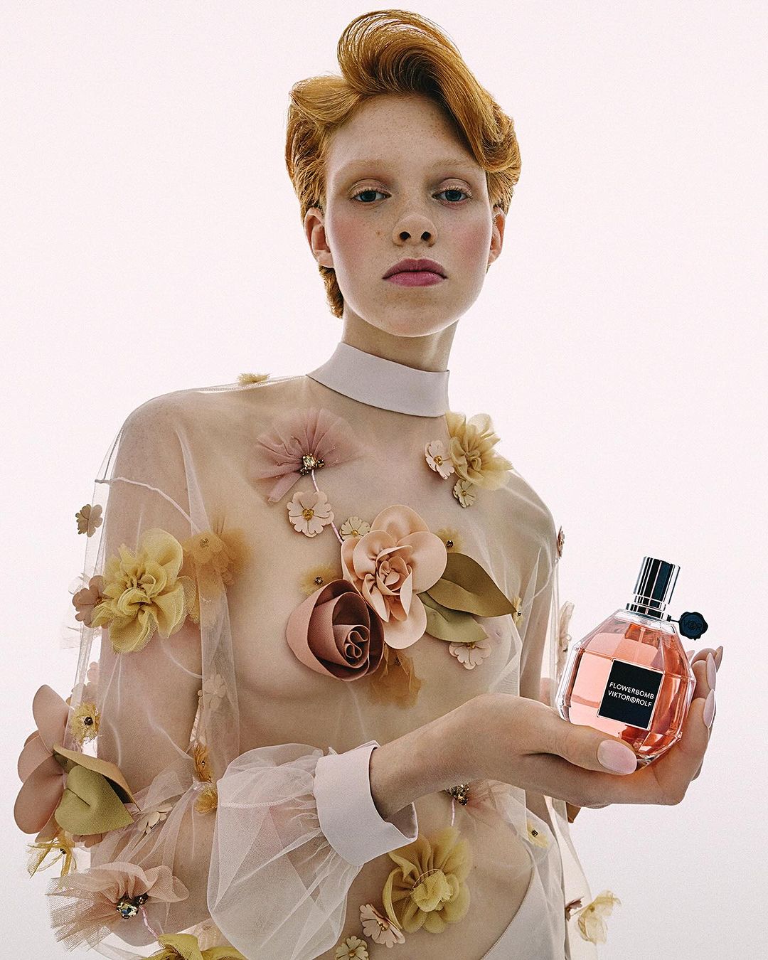 Young female model with red hair poses with a perfume bottle in a fashion shot on a white background with floral top.