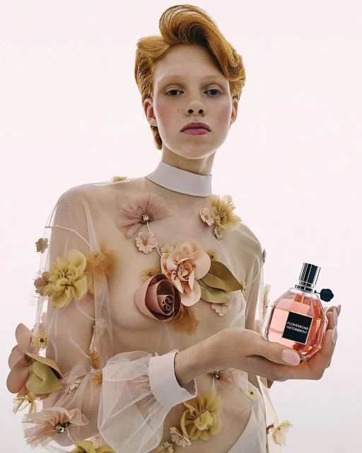 Young female model with red hair poses with a perfume bottle in a fashion shot on a white background with floral top.