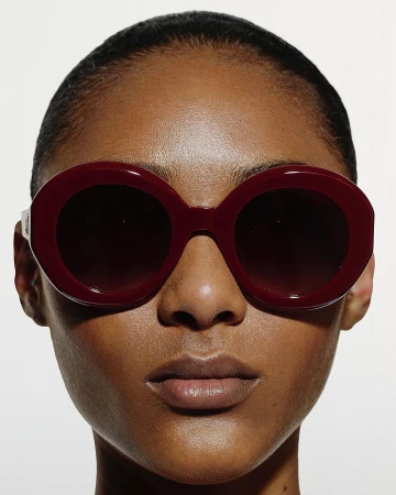 Close-up portrait of a woman wearing oversized burgundy round sunglasses against a plain white background