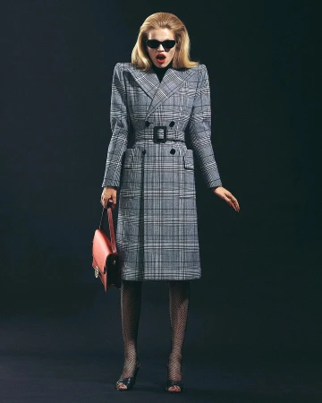 Blonde woman in a structured grey plaid trench coat with large shoulders and black sunglasses, holding a red handbag against a dark backdrop.