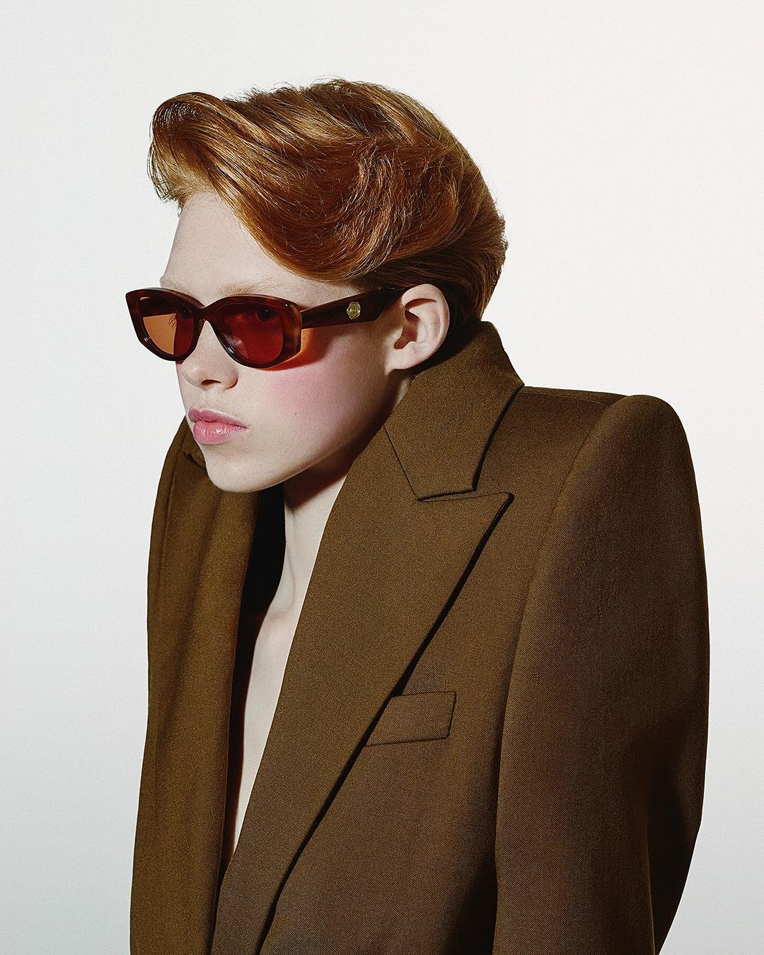 Portrait of red-haired person in sunglasses and brown jacket, standing in a photo studio, shot at eye-level against a white background.