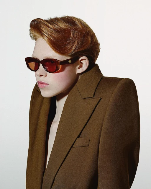 Portrait of red-haired person in sunglasses and brown jacket, standing in a photo studio, shot at eye-level against a white background.