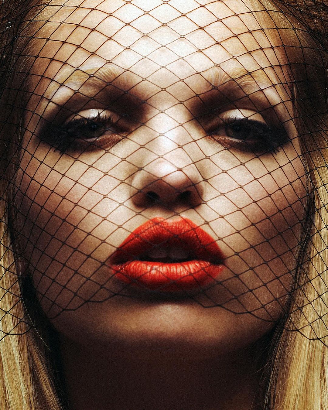 Close-up portrait of a blonde woman with bright red lipstick and a net veil, beauty shot for fashion or cosmetics advertising.