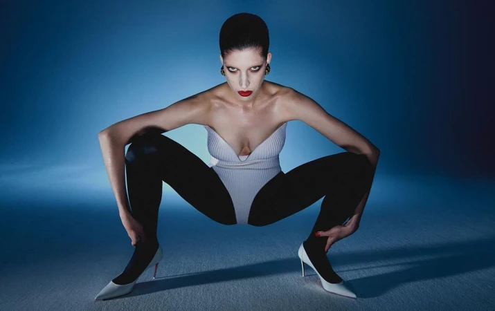 Model in white strapless bodysuit and white heels squatting against a gradient blue studio backdrop by Ferry Van Der Nat.