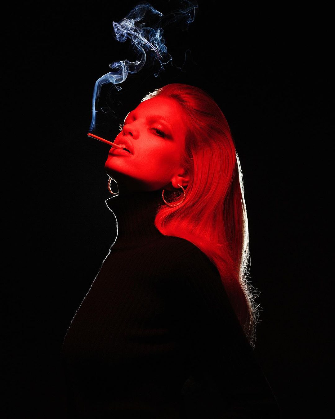 Profile portrait of a woman with long red-lit hair smoking a cigarette against a black background