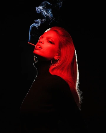 Profile portrait of a woman with long red-lit hair smoking a cigarette against a black background