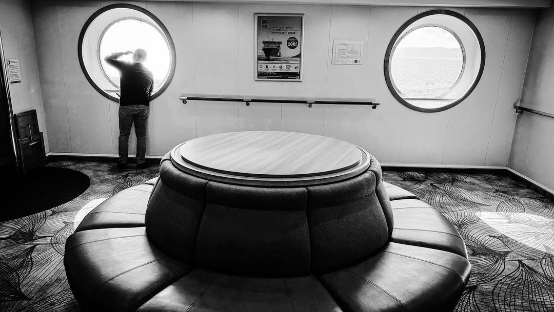 Monochrome interior with a man gazing out a round window on a cruise ship, featuring a circular sofa and patterned carpet.