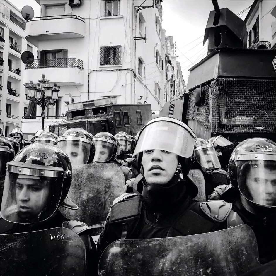 Black and white image of law enforcement officers in riot gear, face forward, with an armored vehicle visible in the background on a city street.