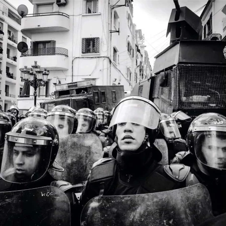 Black and white image of law enforcement officers in riot gear, face forward, with an armored vehicle visible in the background on a city street.