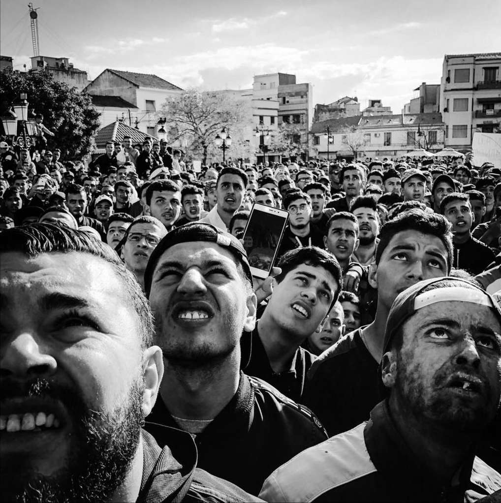 Large crowd of people looking up in an urban square, photographed at eye level in black and white.
