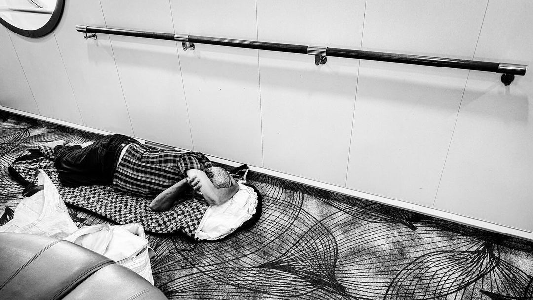 Monochrome image of an adult lying on a patterned blanket on a patterned carpet in an interior setting near a metal handrail.