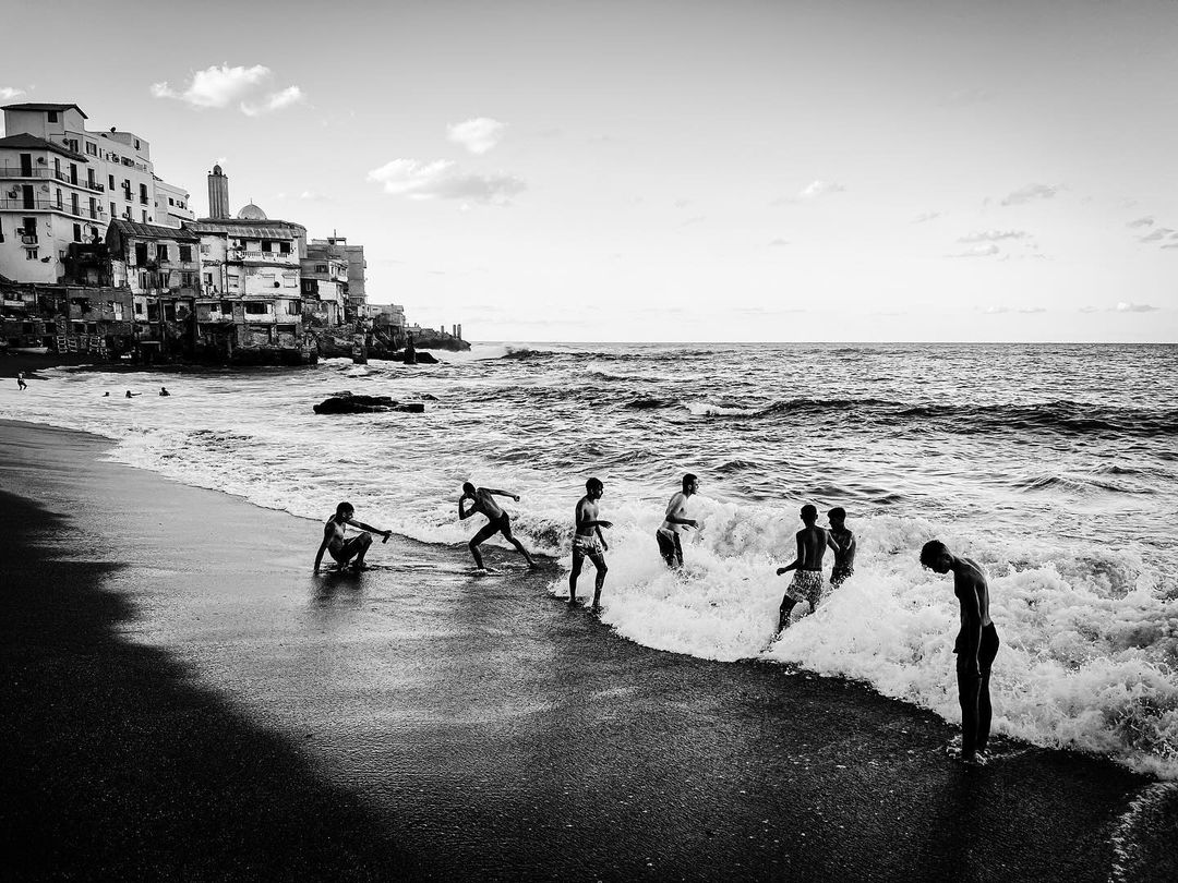 Monochrome image of children playing in the surf on a sandy beach with old buildings in the background under a cloudy sky.