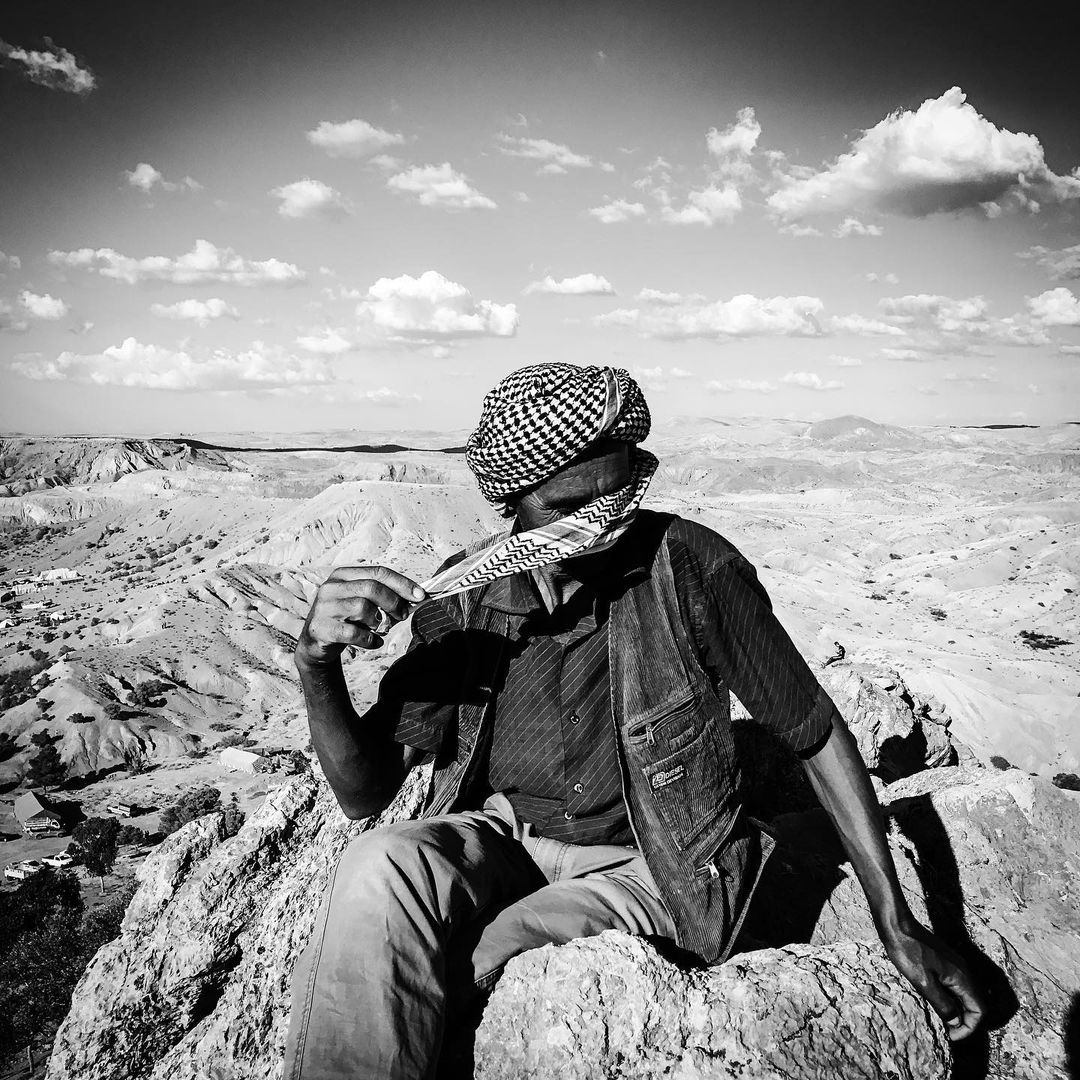 Black and white portrait of a person in a patterned headscarf sitting on a rock in a desert landscape with a cloudy sky above.