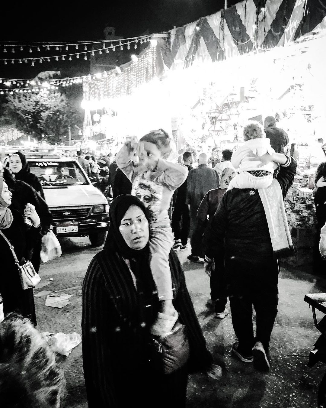 Black and white photo of a woman carrying a child on her shoulders in a crowded outdoor market at night, with a vintage truck nearby.
