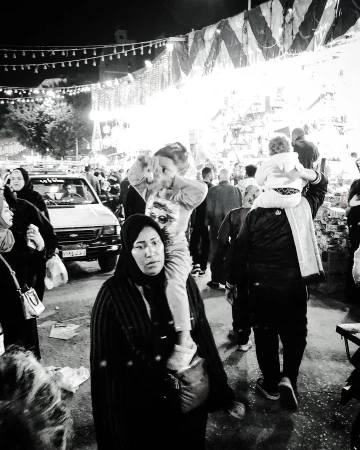 Black and white photo of a woman carrying a child on her shoulders in a crowded outdoor market at night, with a vintage truck nearby.