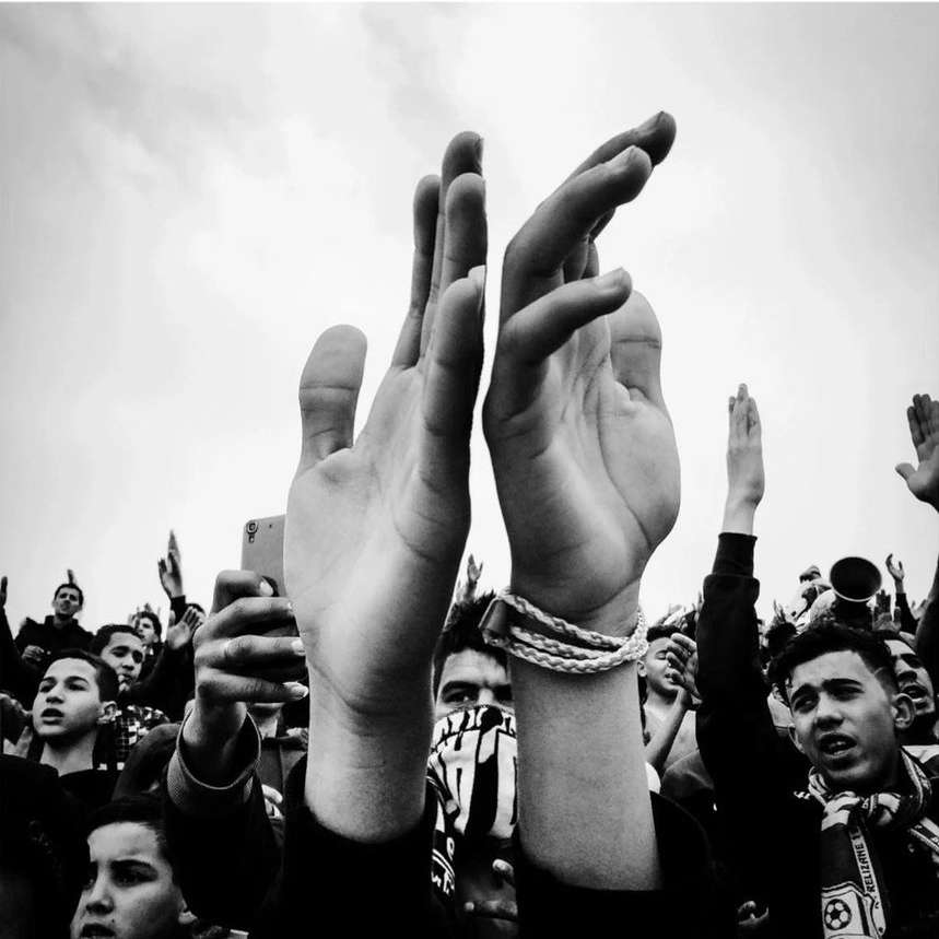 Black and white shot of a large crowd of people cheering outdoors with raised hands and arms, several holding phones to record the scene.