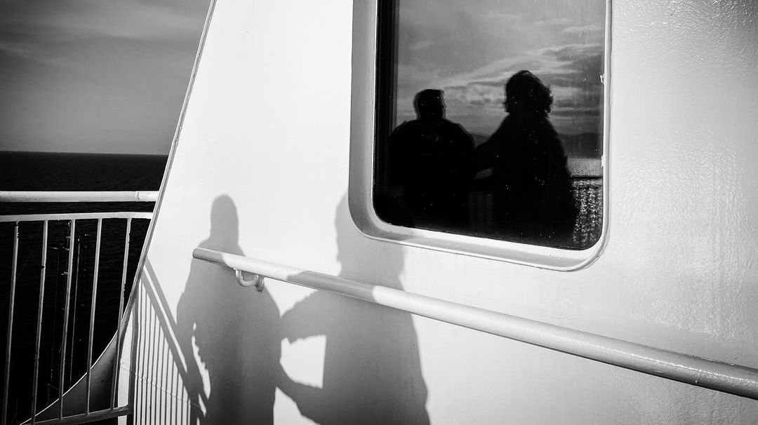 Two figures silhouetted in a window on a boat, monochrome, next to a white wall, rail and a view of the sea in the background.
