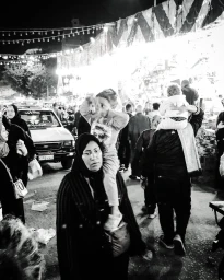 Black and white street photograph of a woman carrying a child on her shoulders in a crowded night market by Fethi Sahraoui