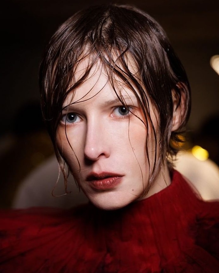 Close-up portrait of a young woman with wet brown hair, light eyes, and dark red lipstick, wearing a red top. Dark, blurred background.