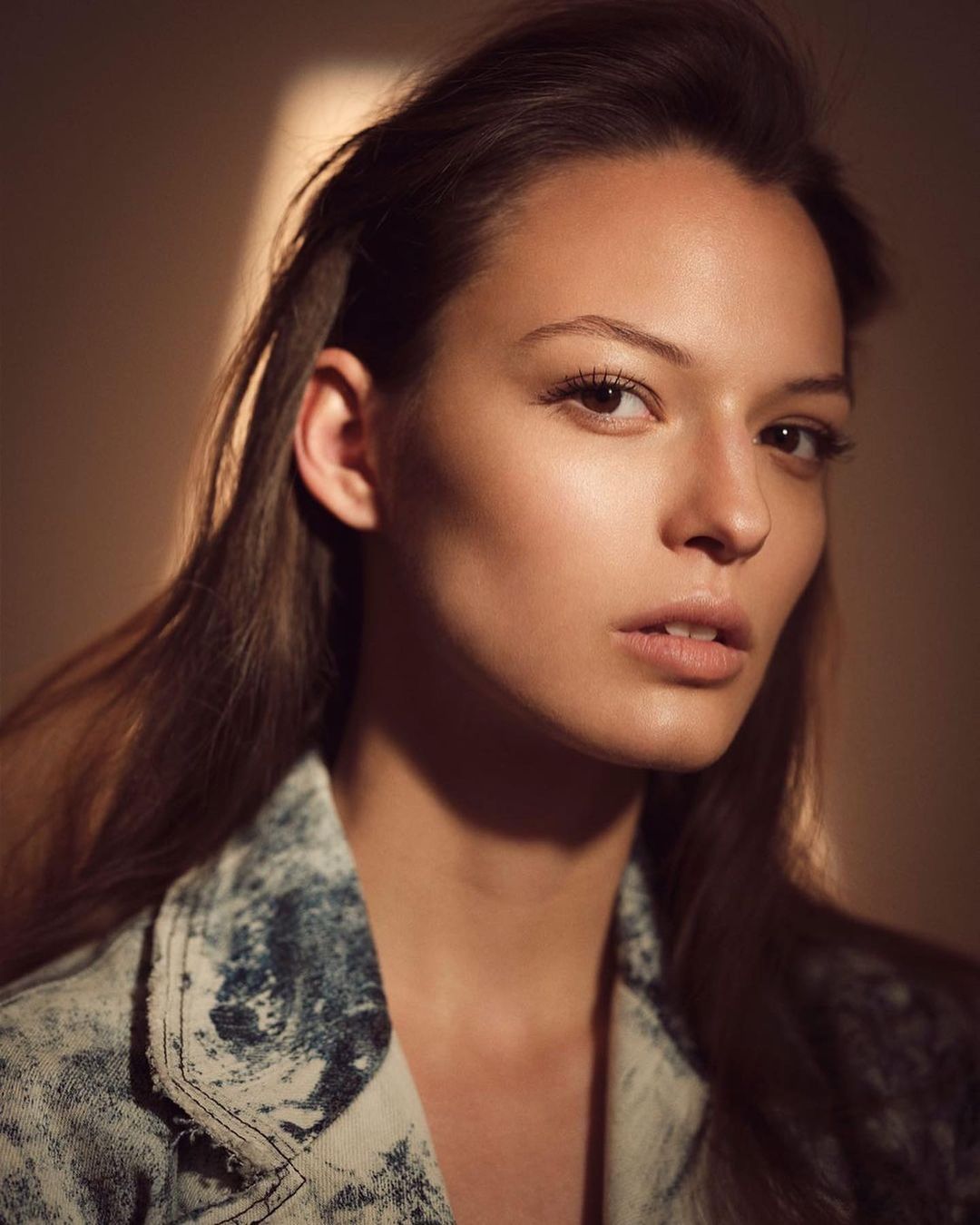 Portrait of a young woman with long brown hair wearing a blue denim jacket in front of a blurred tan background, illuminated by window light.