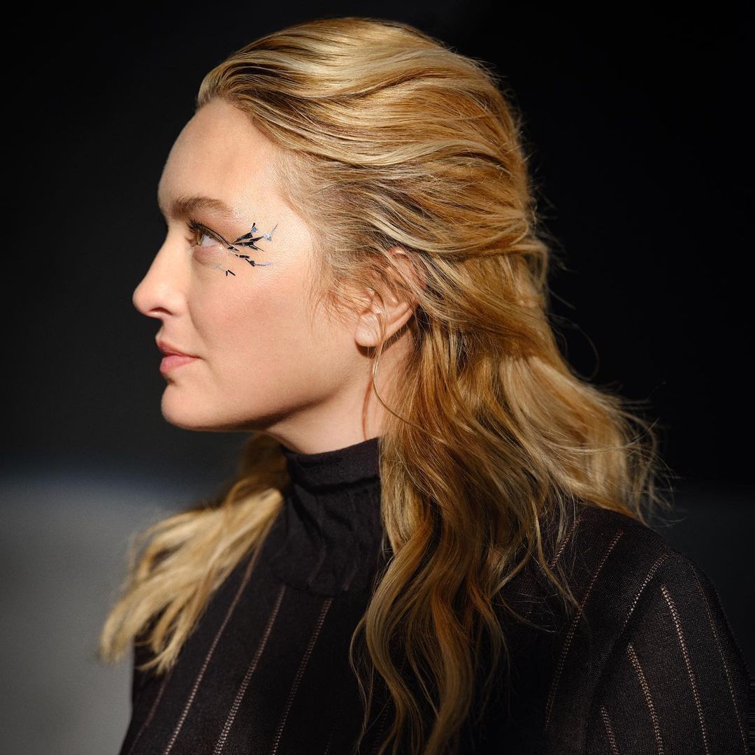 Profile view of a blonde woman with creative makeup and a black pinstripe top against a dark, blurred background in a studio setting.