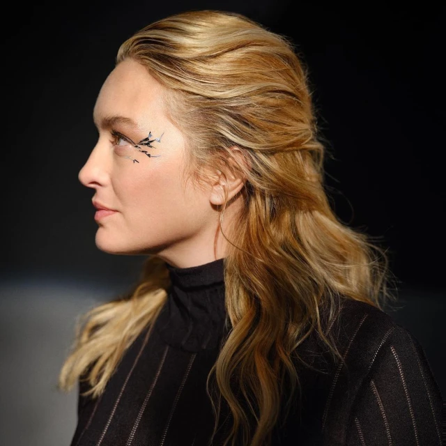 Profile view of a blonde woman with creative makeup and a black pinstripe top against a dark, blurred background in a studio setting.