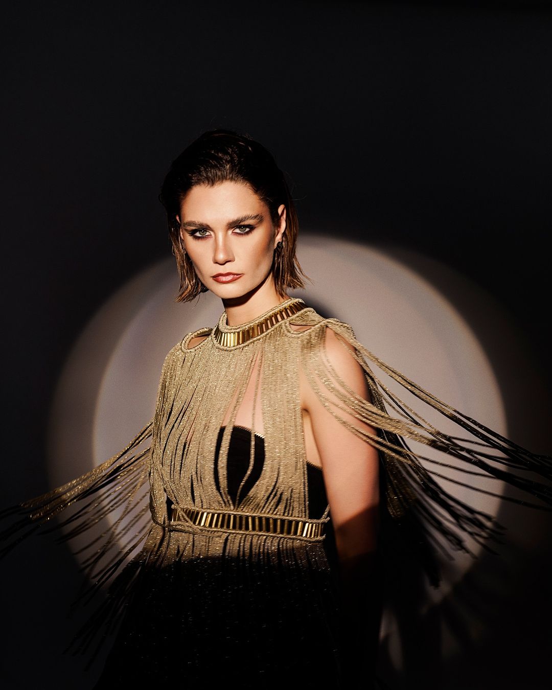 An adult woman with short brown hair is posed in front of a spotlight wearing a gold fringe dress with a black panel and gold collar.