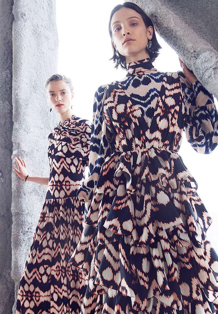 Two fair-skinned women wearing black and beige patterned tiered dresses stand amidst stone ruins in a fashion editorial style photograph.