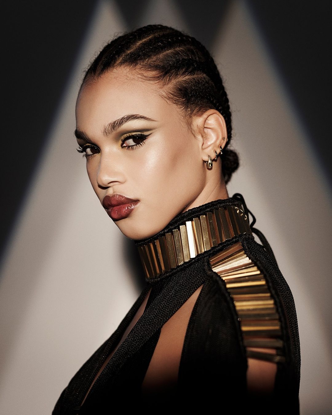 A striking close-up captures a woman with braided hair, glossy lips, and green eye makeup, wearing a black top with a gold collar.