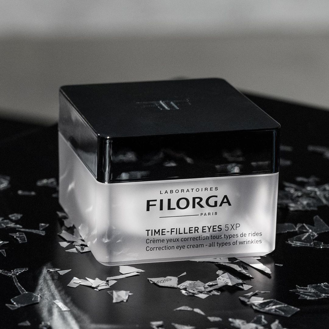 Close-up of Filorga Time-Filler Eyes 5XP eye cream jar with black lid and silver accents, set against a dark background with silver shards.