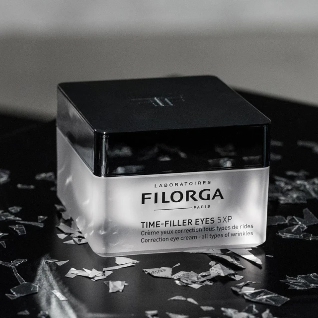 Close-up of Filorga Time-Filler Eyes 5XP eye cream jar with black lid and silver accents, set against a dark background with silver shards.