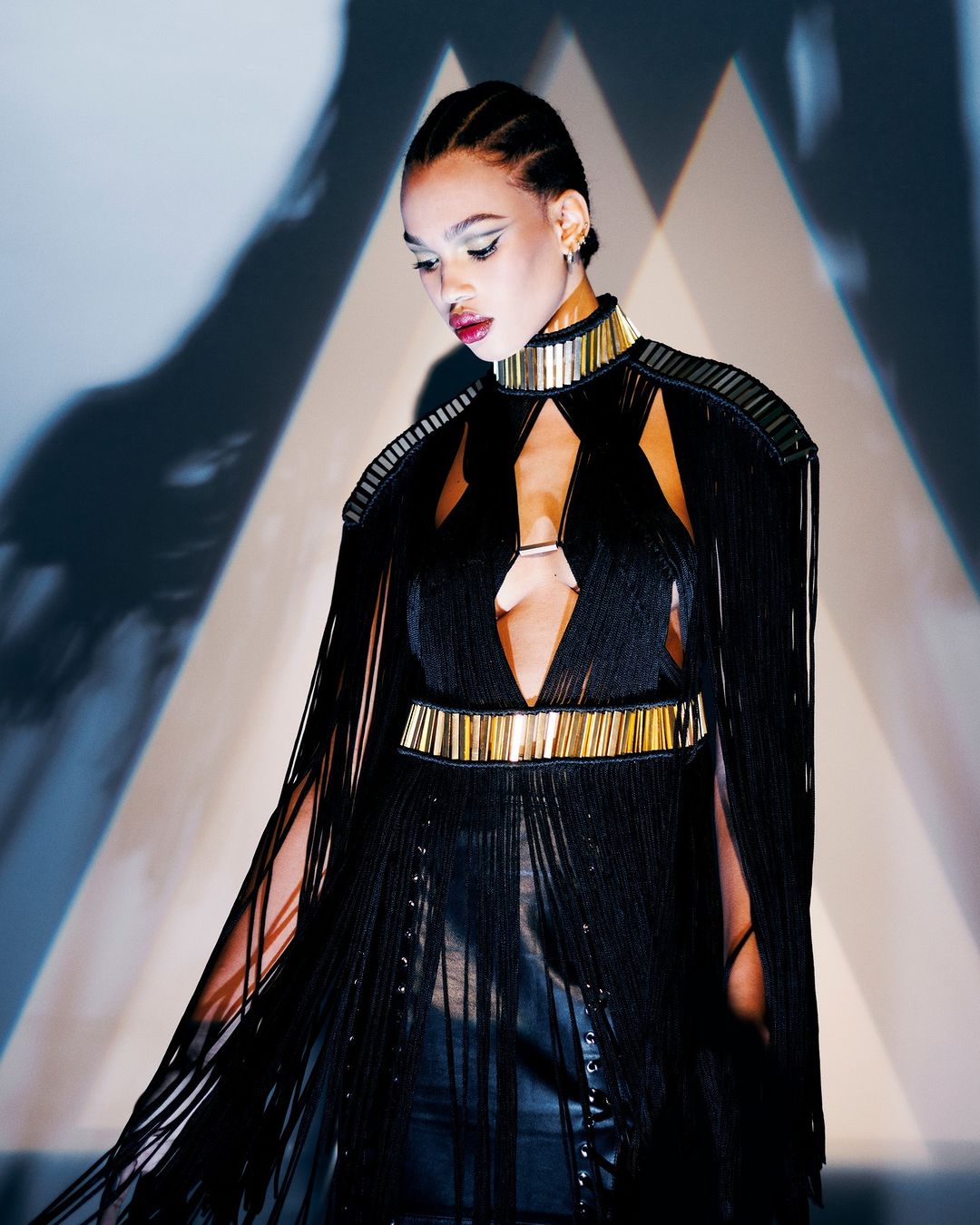 Woman in black fringe dress with gold accents poses in a photo studio against a geometric backdrop with light and shadow play.