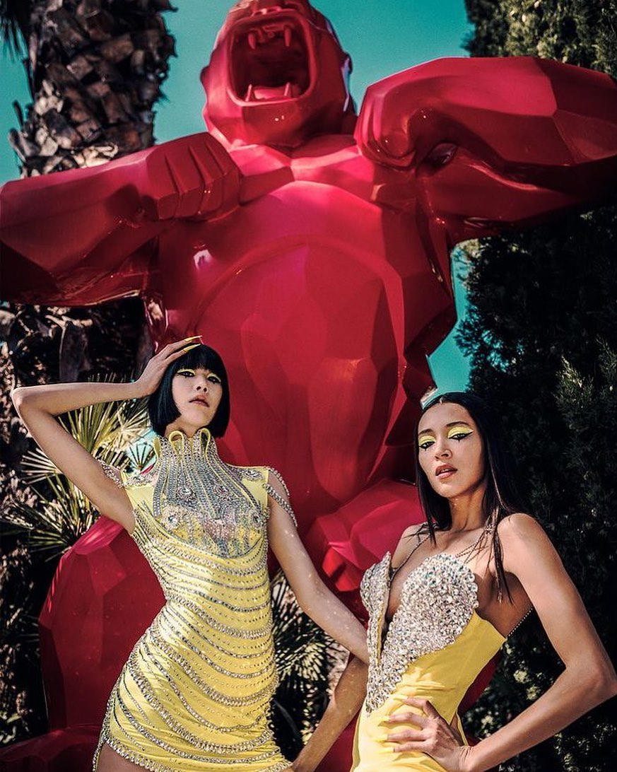 Two women in yellow dresses pose near a large red gorilla statue outdoors on a sunny day, showcasing fashion and modern sculpture.