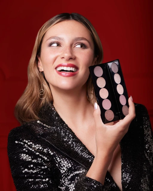 Smiling woman holds eyeshadow palette in studio portrait against red background. She wears black sequin jacket and gold earrings.