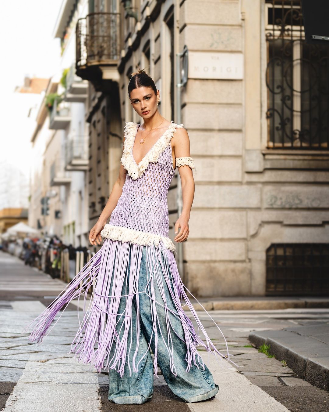 A woman poses in a purple crochet dress with fringe and wide-leg jeans on an urban street in daylight, fashion editorial photography.