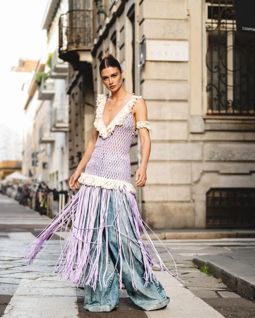 A woman poses in a purple crochet dress with fringe and wide-leg jeans on an urban street in daylight, fashion editorial photography.