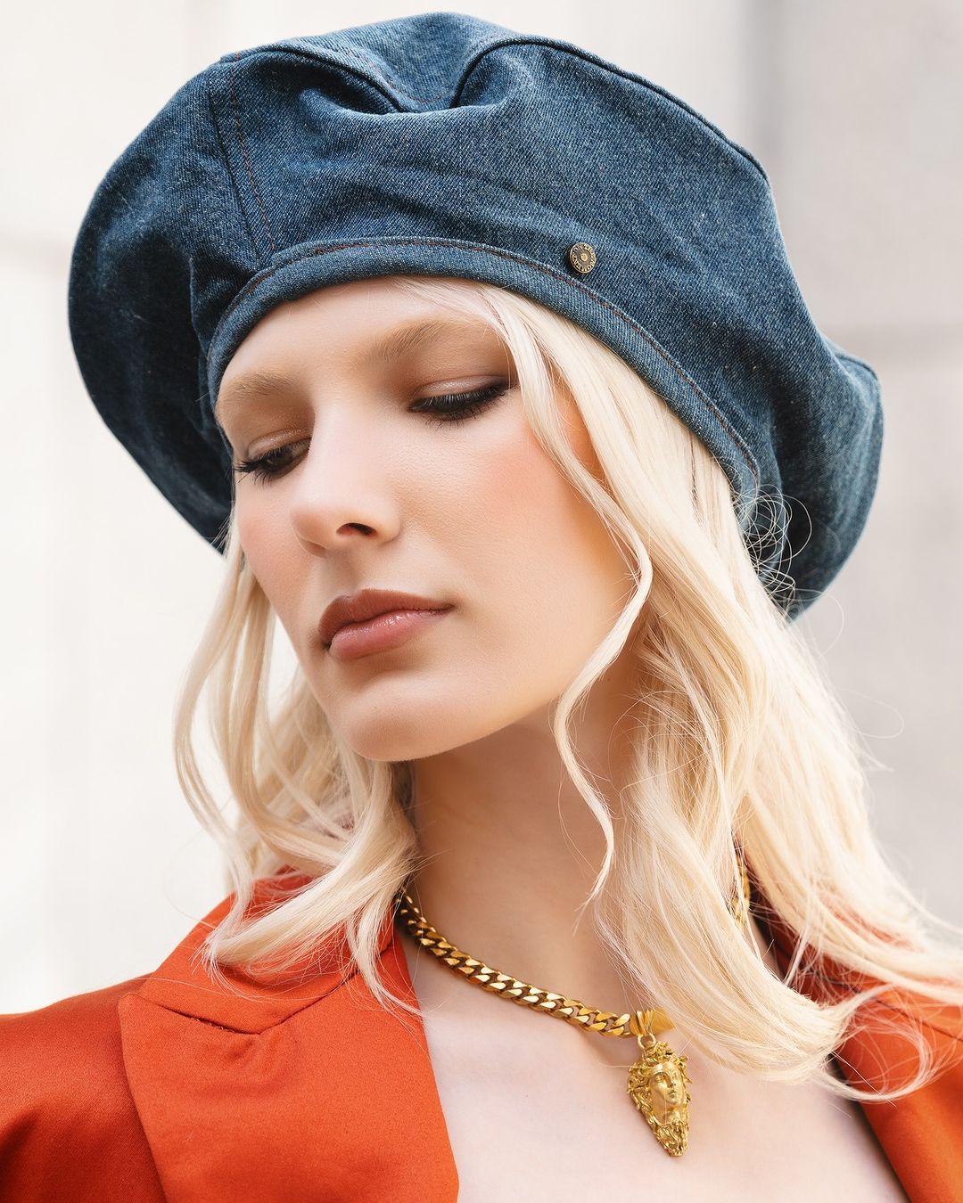 Portrait of a woman with blonde hair wearing a blue denim beret and an orange blazer with a gold chain necklace and pendant.